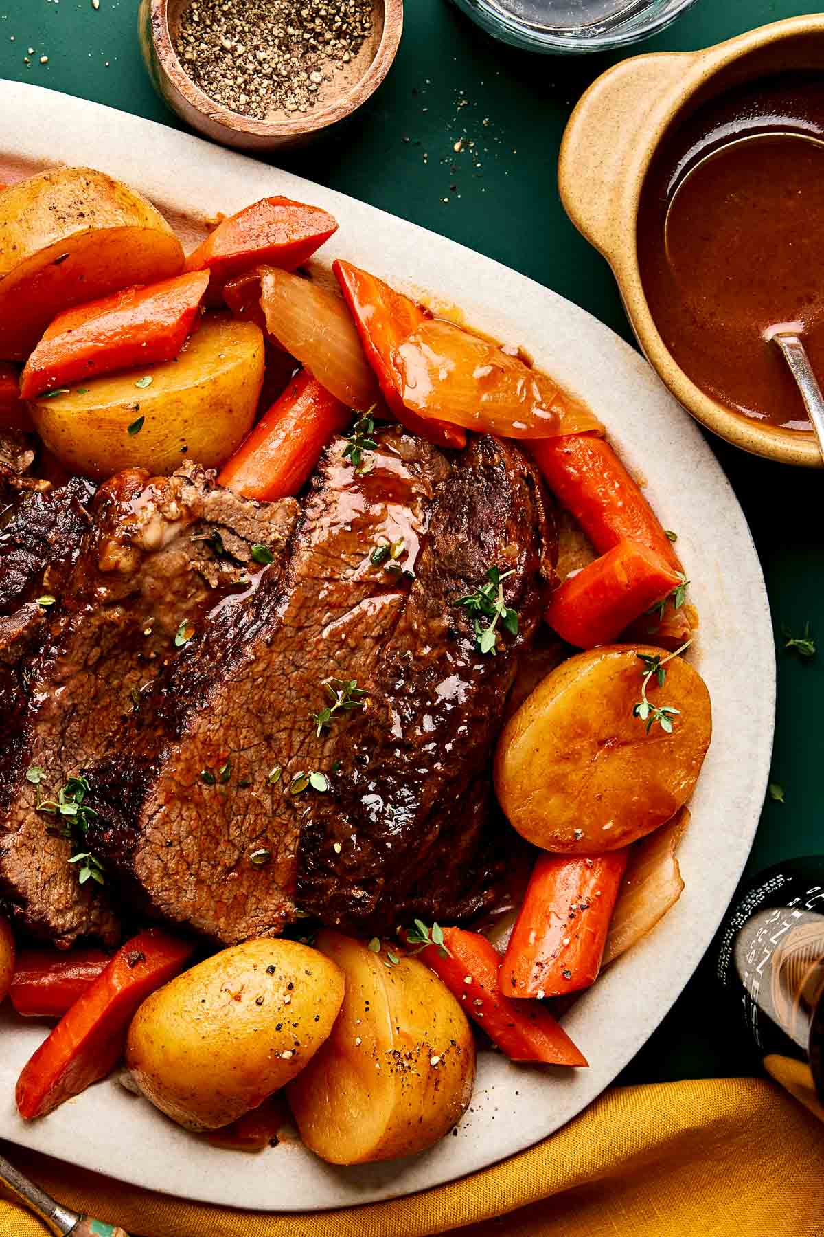 A platter of pot roast with sliced beef, potatoes, carrots, and onions, garnished with fresh herbs. A bowl of brown gravy and a small dish of ground black pepper are nearby.