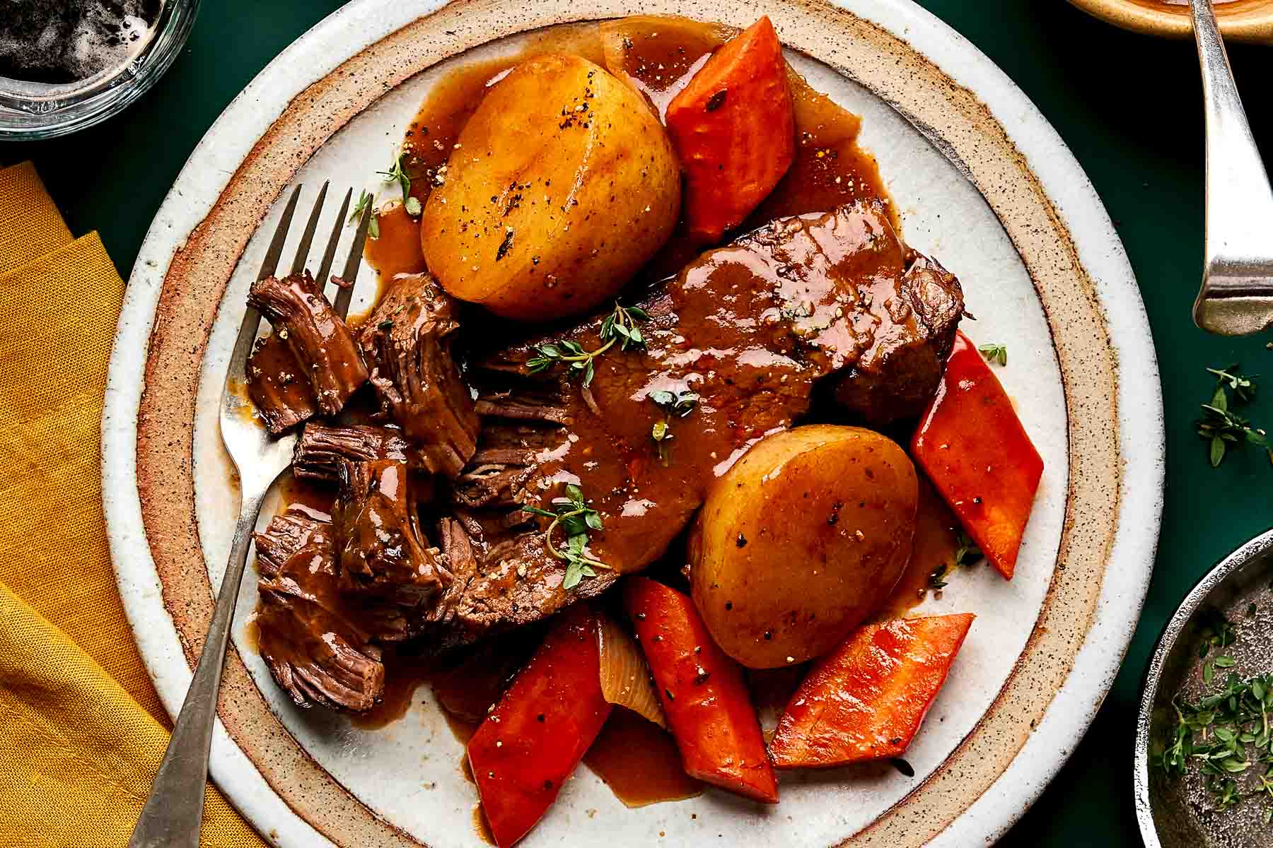 A plate of pot roast with gravy, served with potatoes and carrots. A fork is shredding some of the tender beef. The dish is garnished with fresh herbs and set on a rustic white plate.