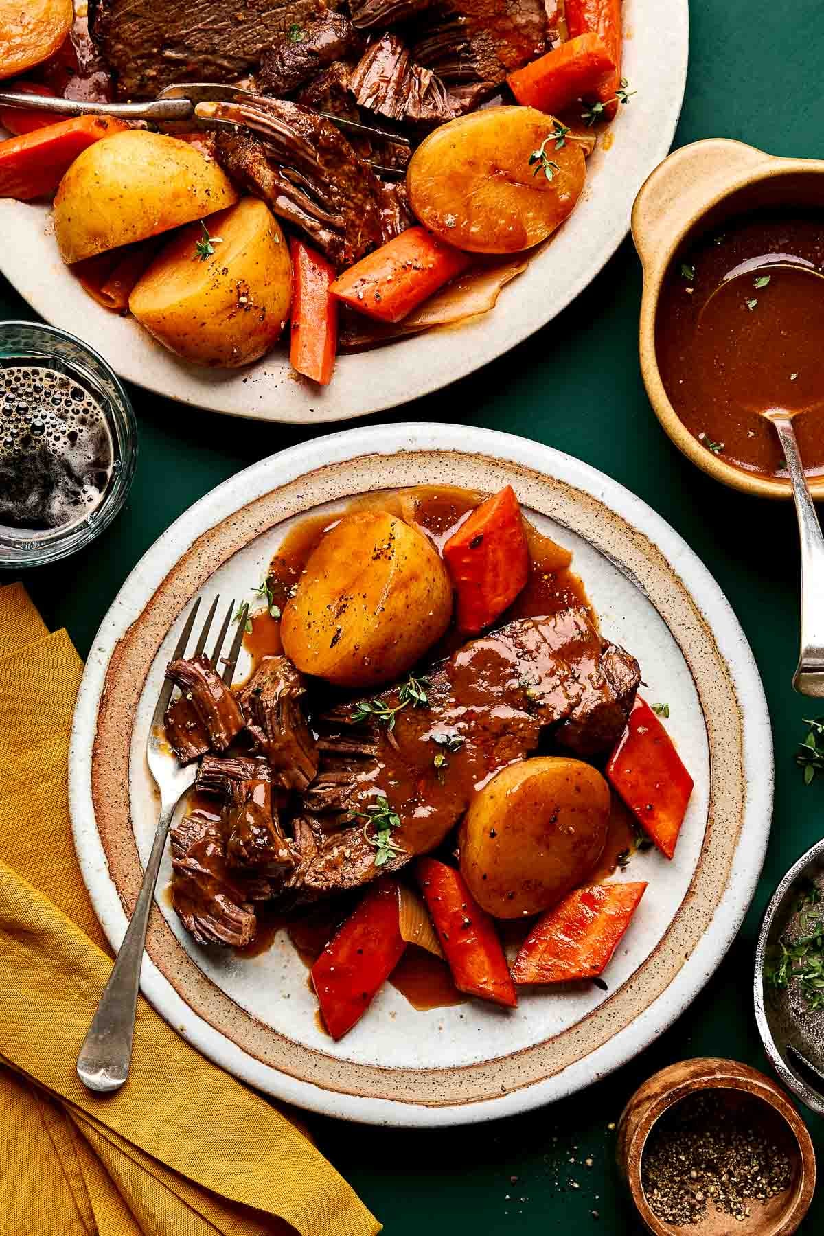 Plated Guinness Pot Roast A plate of pot roast with gravy, served with potatoes and carrots, sits on a table next to a bowl of extra gravy, a glass of dark beverage, and a napkin with utensils.