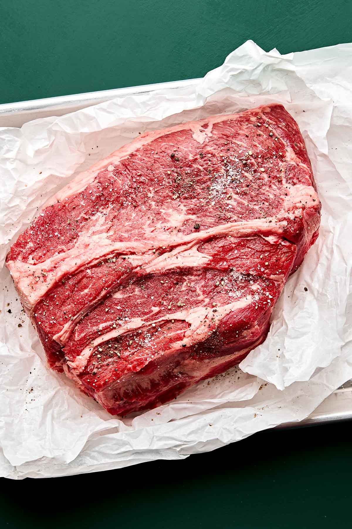 A large, raw beef roast seasoned with salt and pepper rests on white parchment paper atop a metal baking sheet, set against a green background.