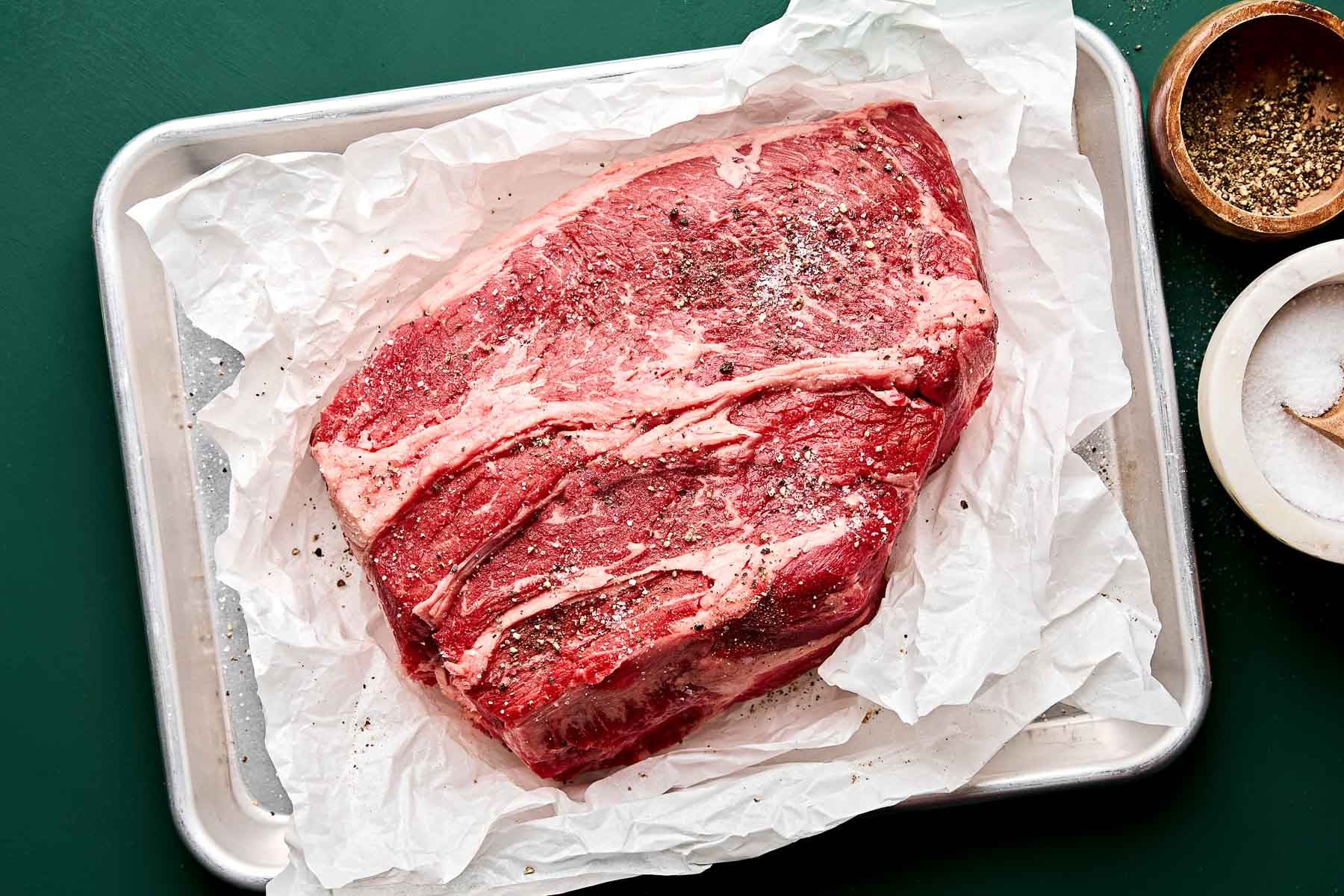 A raw, marbled cut of beef brisket seasoned with pepper and salt sits on white parchment paper atop a baking sheet, with bowls of salt and pepper visible nearby.