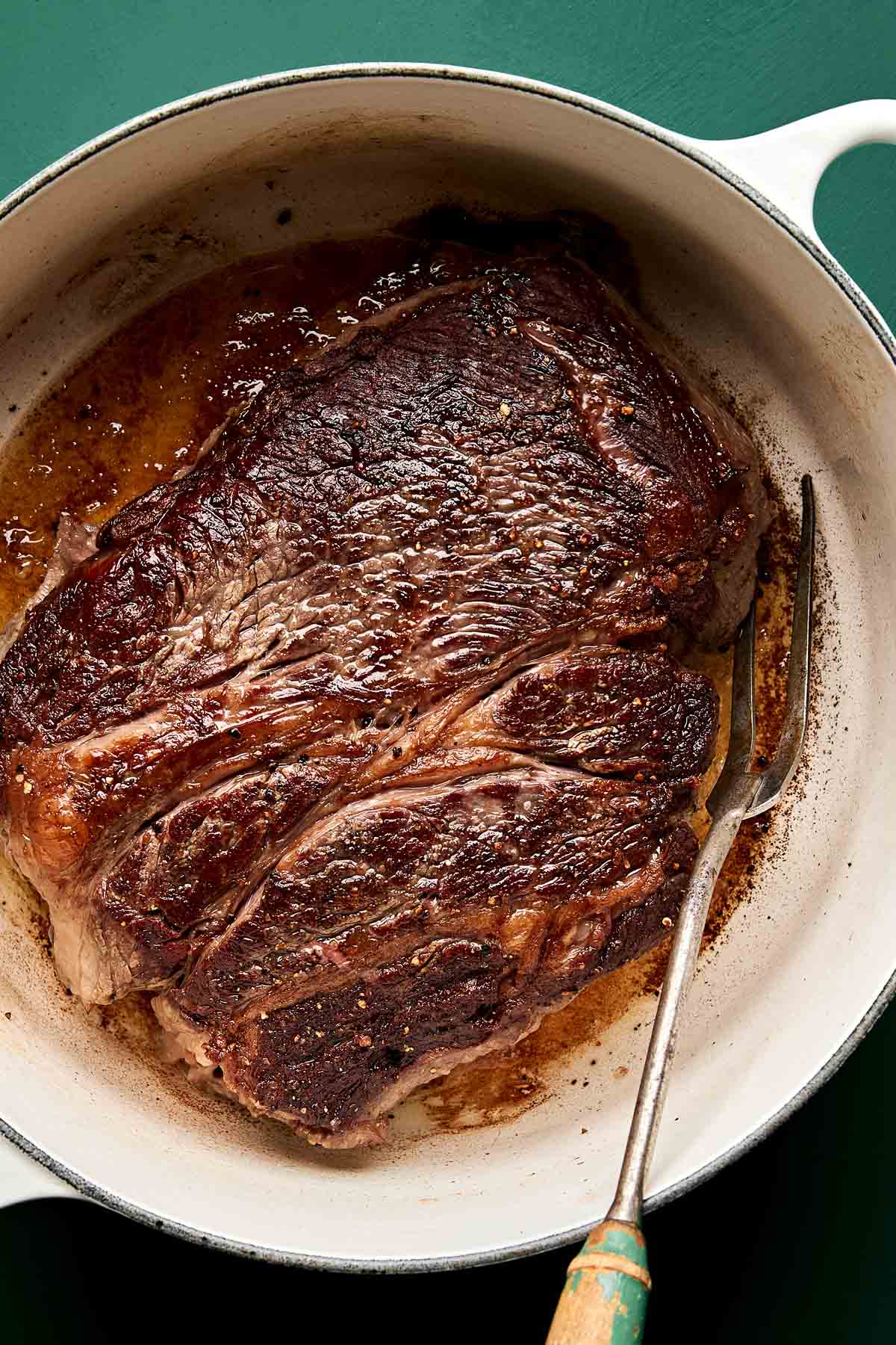 Searing chuck roast A large, browned roast sits in a white Dutch oven with juices around it. A two-pronged meat fork rests beside the roast on the right. The background is a dark green surface.