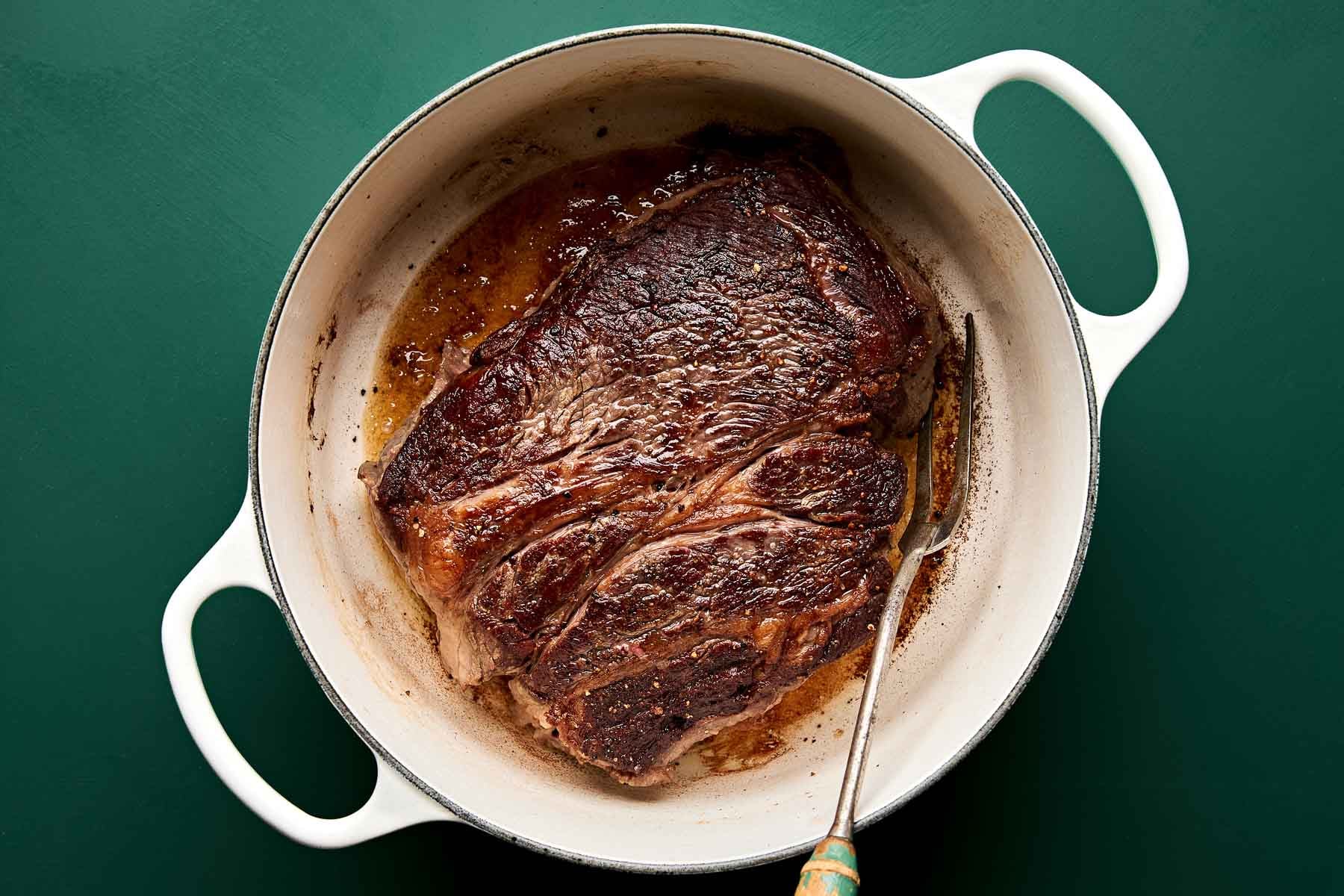 A browned, cooked beef roast sits in a white Dutch oven with some juices, against a dark green background. A meat fork rests next to the roast inside the pot.
