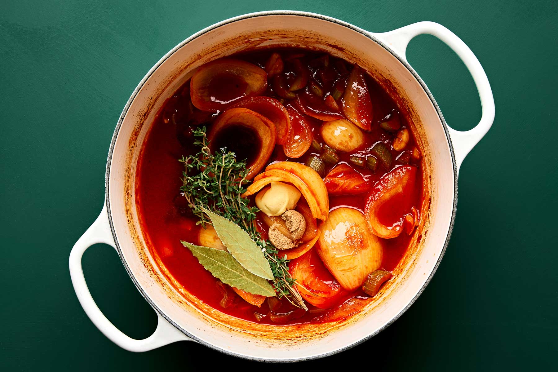 A white pot filled with a tomato-based stew featuring onions, garlic, tomatoes, mushrooms, celery, herbs like thyme, and bay leaves, on a dark green surface.