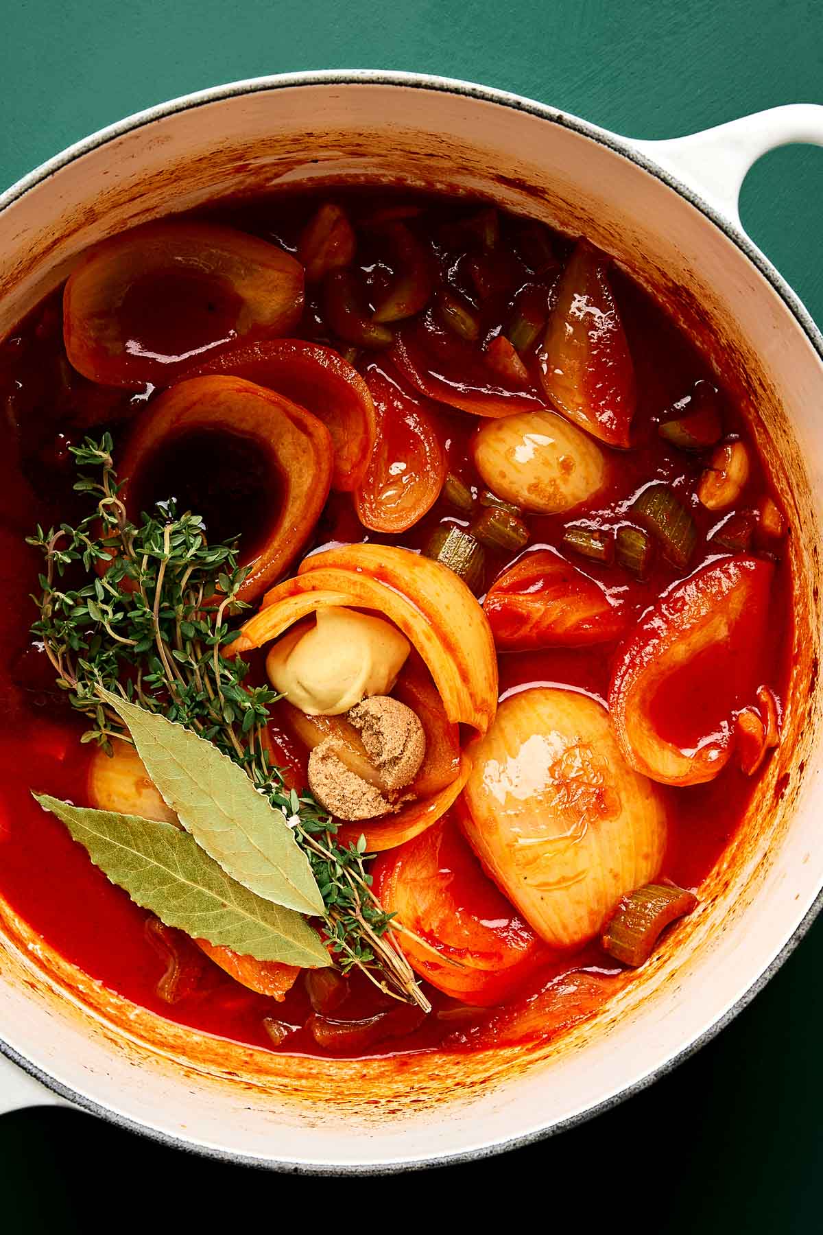 Aromatic base for pot roast A white pot filled with tomato-based stew containing onions, celery, tomatoes, fresh thyme, bay leaves, garlic cloves, and spices, set against a dark green background.