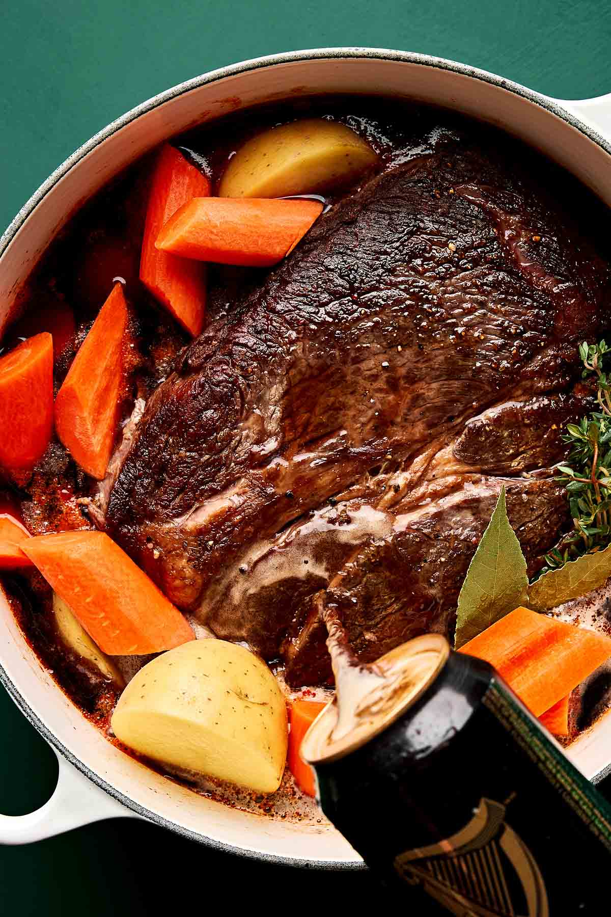 Pouring Beer Into Pot Roast A large piece of beef roast in a pot with cut potatoes, carrots, herbs, and bay leaves, as a can of Guinness stout is being poured over the ingredients.