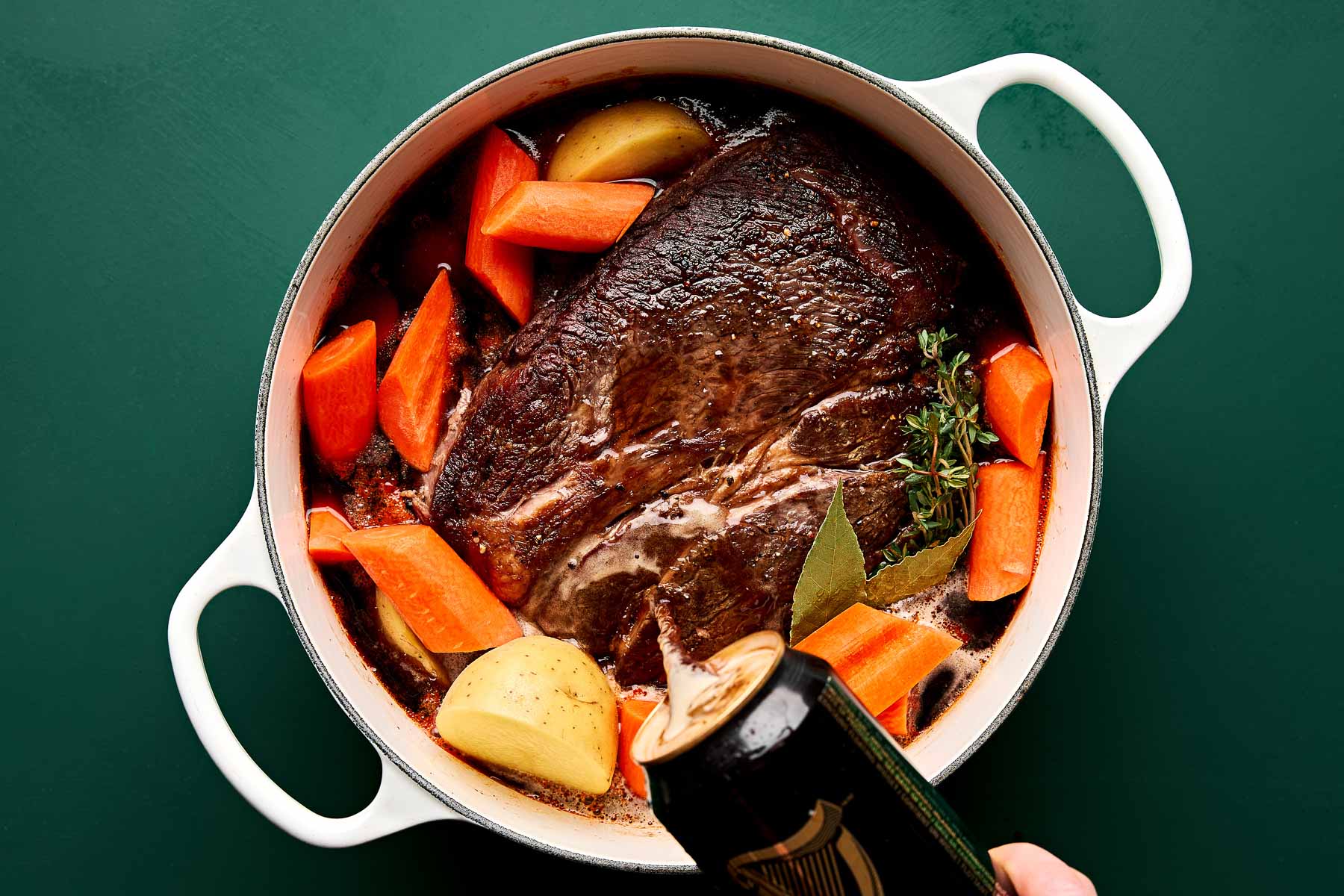 A white Dutch oven containing a cooked pot roast with carrots, potatoes, herbs, and a hand pouring a can of dark beer over the meat, set against a dark green background.