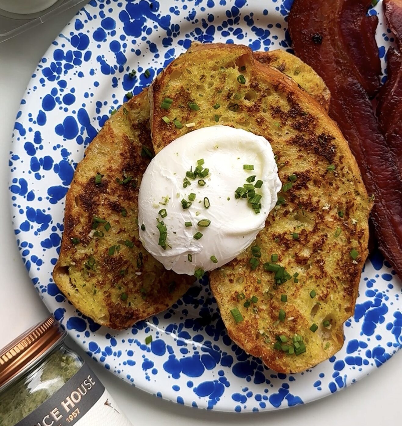 A blue speckled plate with two slices of herb toast topped with a poached egg and chopped chives, served with two strips of bacon. A spice jar and eggs in a carton are nearby on a white surface.