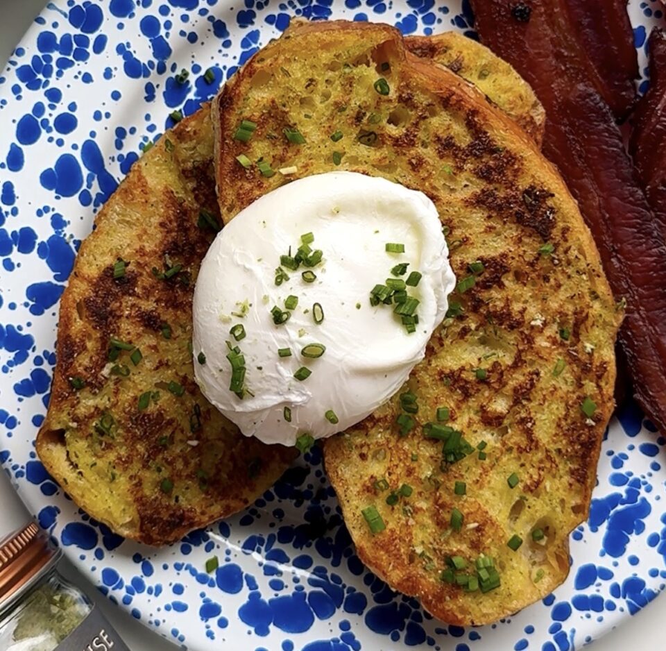 A plate with two slices of herb toast topped with a poached egg and chives, served with two strips of bacon. Fresh eggs and a jar of shallot & herb seasoning are nearby. The plate has a blue splatter pattern.