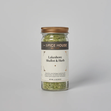 A glass jar of The Spice House Lakeshore Shallot & Herb seasoning, featuring a brown and white label with gold accents, filled with a green herb blend, set against a plain gray background.