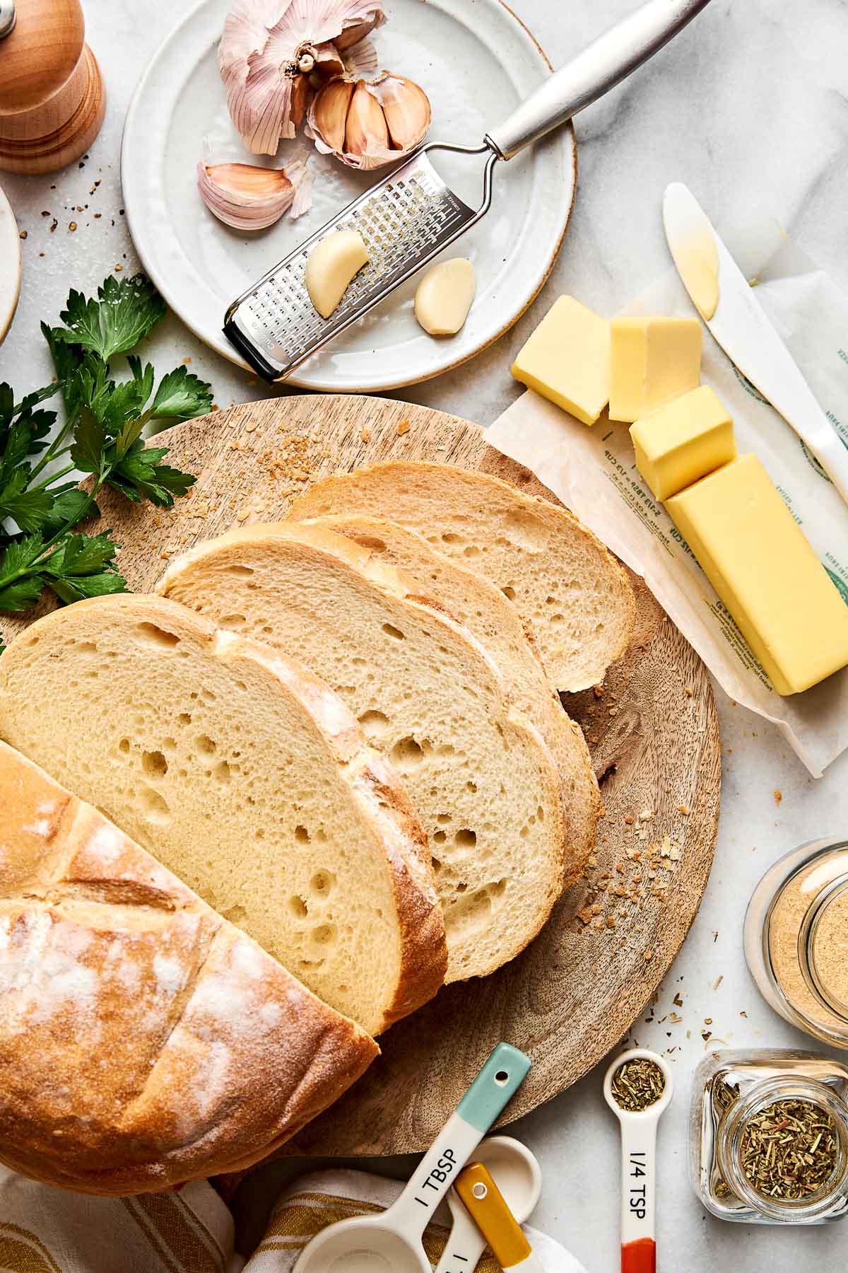 A round loaf of bread, partially sliced, sits on a wooden board. Nearby are butter sticks, garlic cloves, a grater, fresh parsley, measuring spoons, and herbs on a white countertop.
