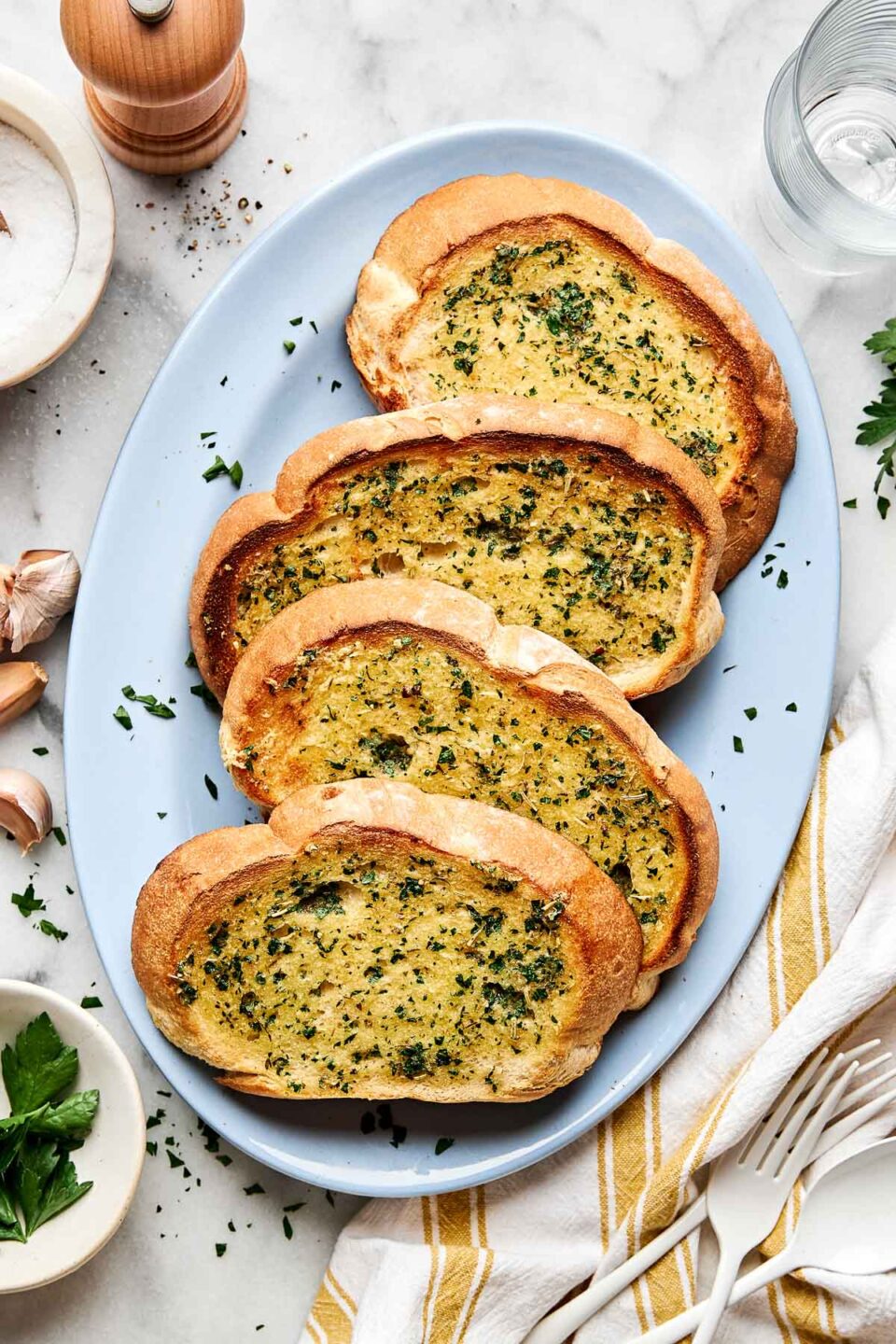 Four slices of golden-brown garlic bread topped with chopped parsley are arranged on a light blue oval plate, set on a table with garlic cloves, parsley, salt, a pepper grinder, and utensils nearby.