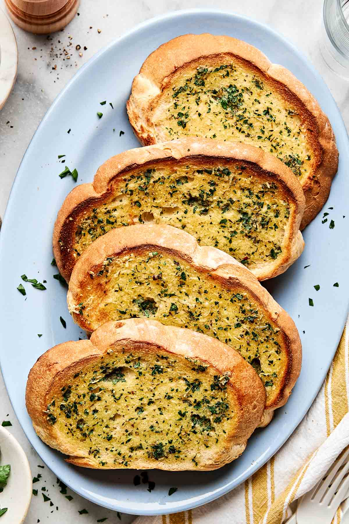 A blue plate with four slices of toasted garlic bread, topped with chopped herbs and garlic butter, set on a white surface with scattered parsley.