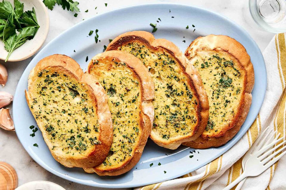Four slices of toasted garlic bread with chopped parsley on top are arranged on a light blue oval plate, with a few sprigs of parsley and a garlic clove nearby.