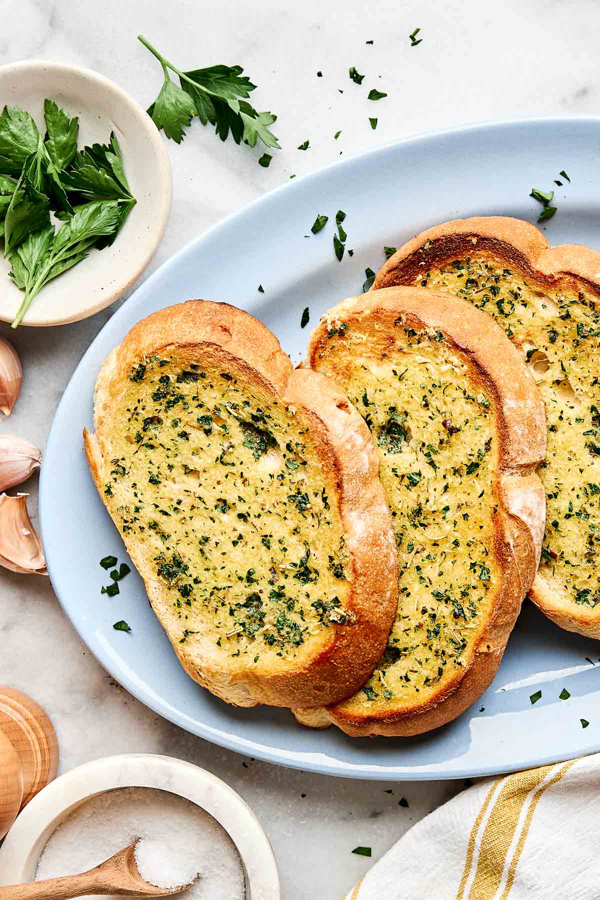 Three slices of garlic bread with herbs on a white plate, surrounded by fresh parsley, garlic cloves, and a wooden salt cellar on a marble surface.