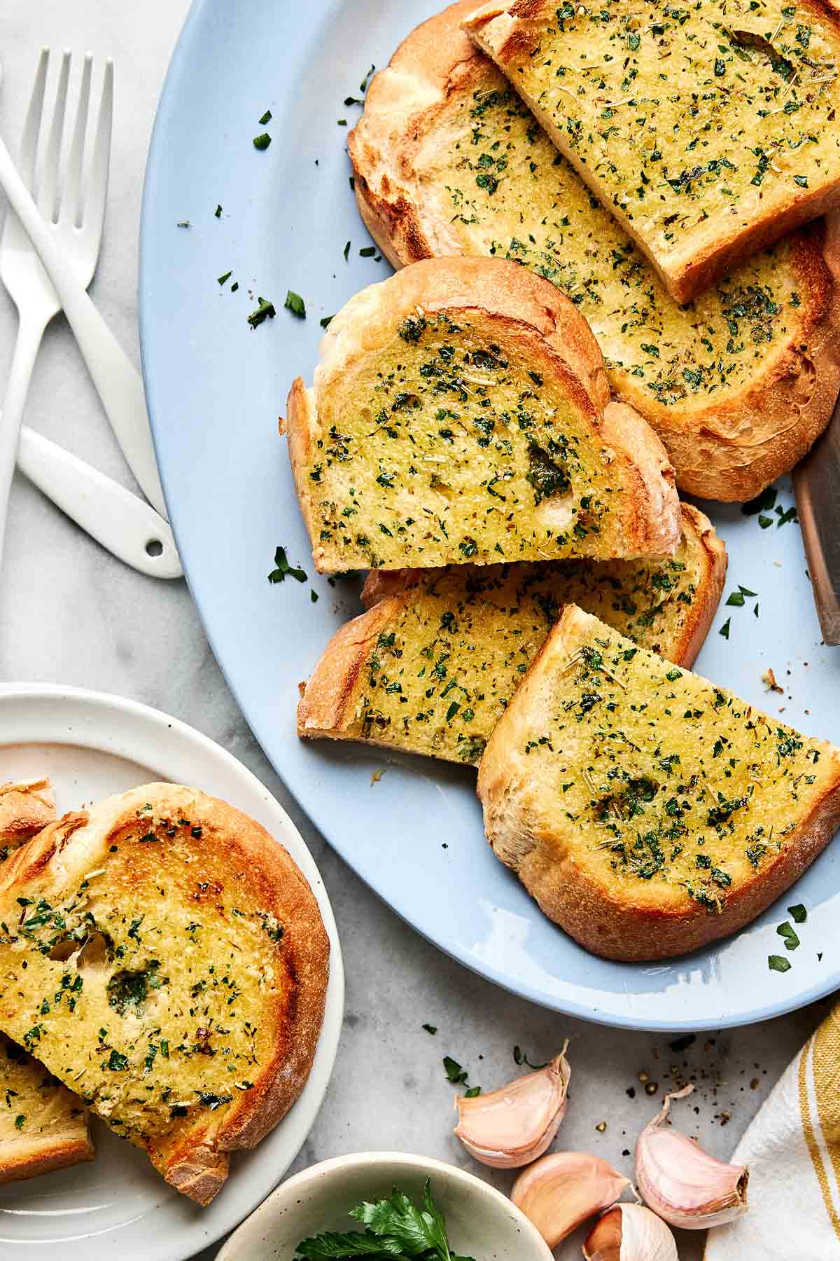 Slices of golden, toasted garlic bread topped with chopped herbs are arranged on a light blue platter, with a few slices on a small white plate beside it. Garlic cloves and utensils are nearby on a marble surface.