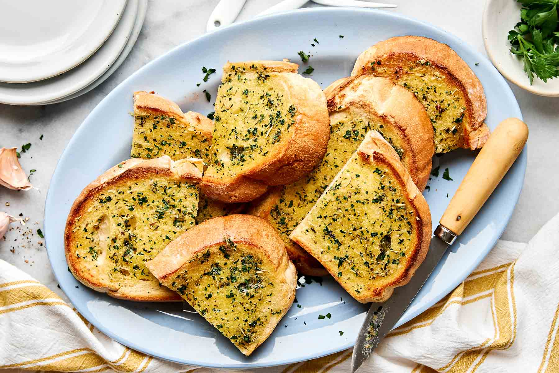 A blue oval plate holds several slices of golden, crispy garlic bread sprinkled with chopped parsley. A knife rests on the plate, and a bowl of fresh herbs and a striped towel are nearby.