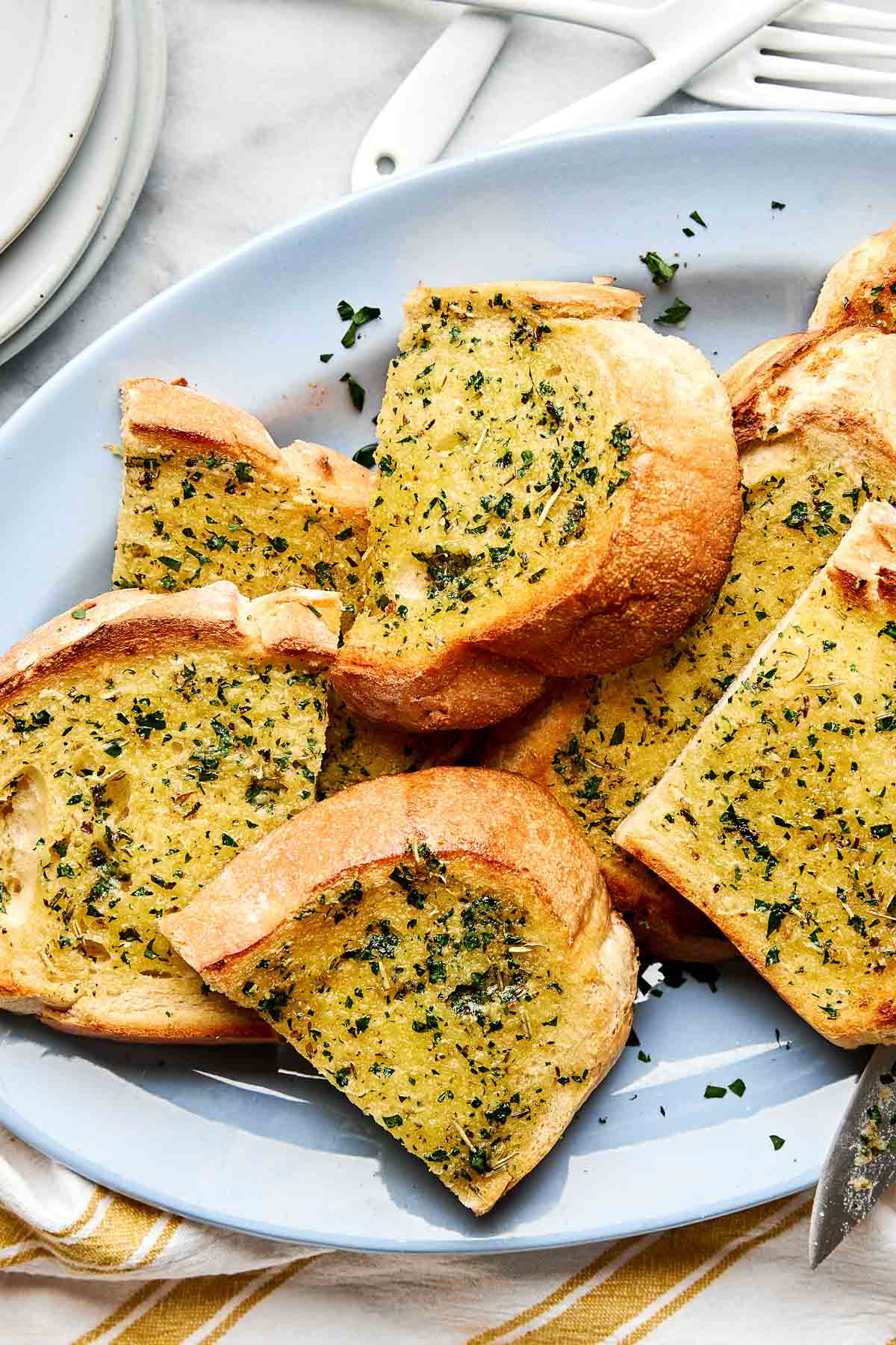 Several slices of golden brown garlic bread topped with chopped herbs are arranged on a light blue plate, placed on a white and yellow striped cloth next to some plates and utensils.