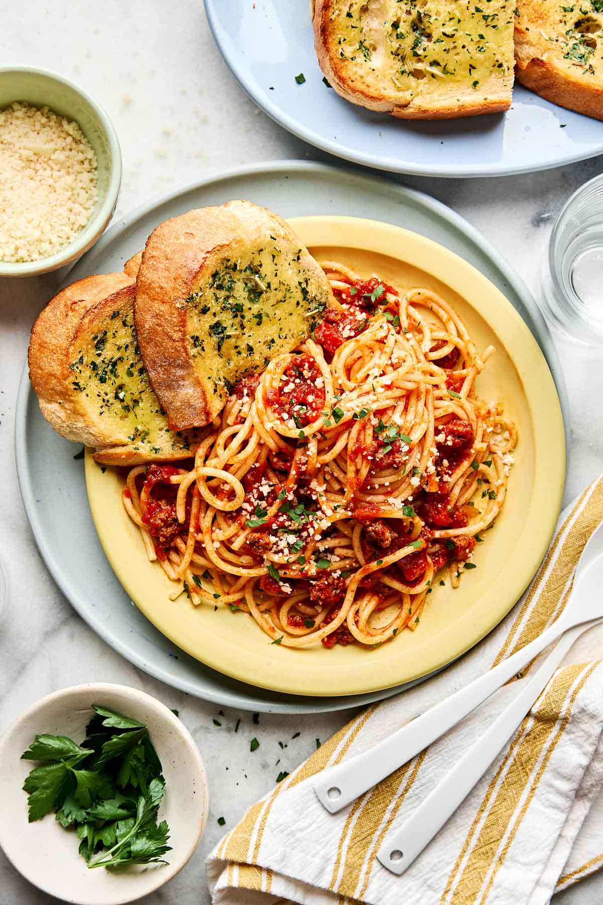 Plate of spaghetti with tomato sauce, grated cheese, and herbs, served with two slices of garlic bread. Nearby are a bowl of grated cheese, fresh parsley, a plate of more garlic bread, and utensils on a striped napkin.