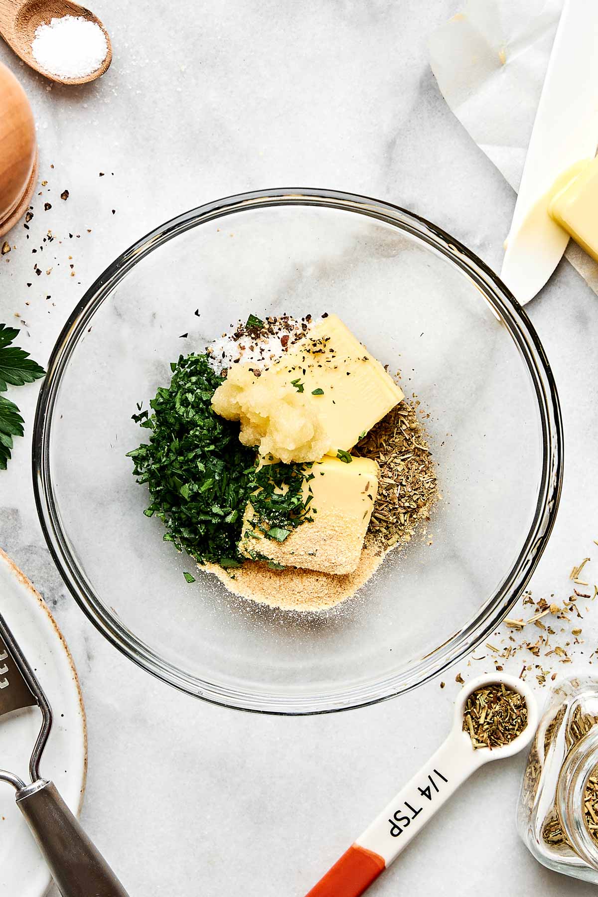 A clear glass bowl contains butter, chopped parsley, minced garlic, dried herbs, and breadcrumbs on a marble countertop, surrounded by an egg, parsley, a grater, a knife, and a measuring spoon.