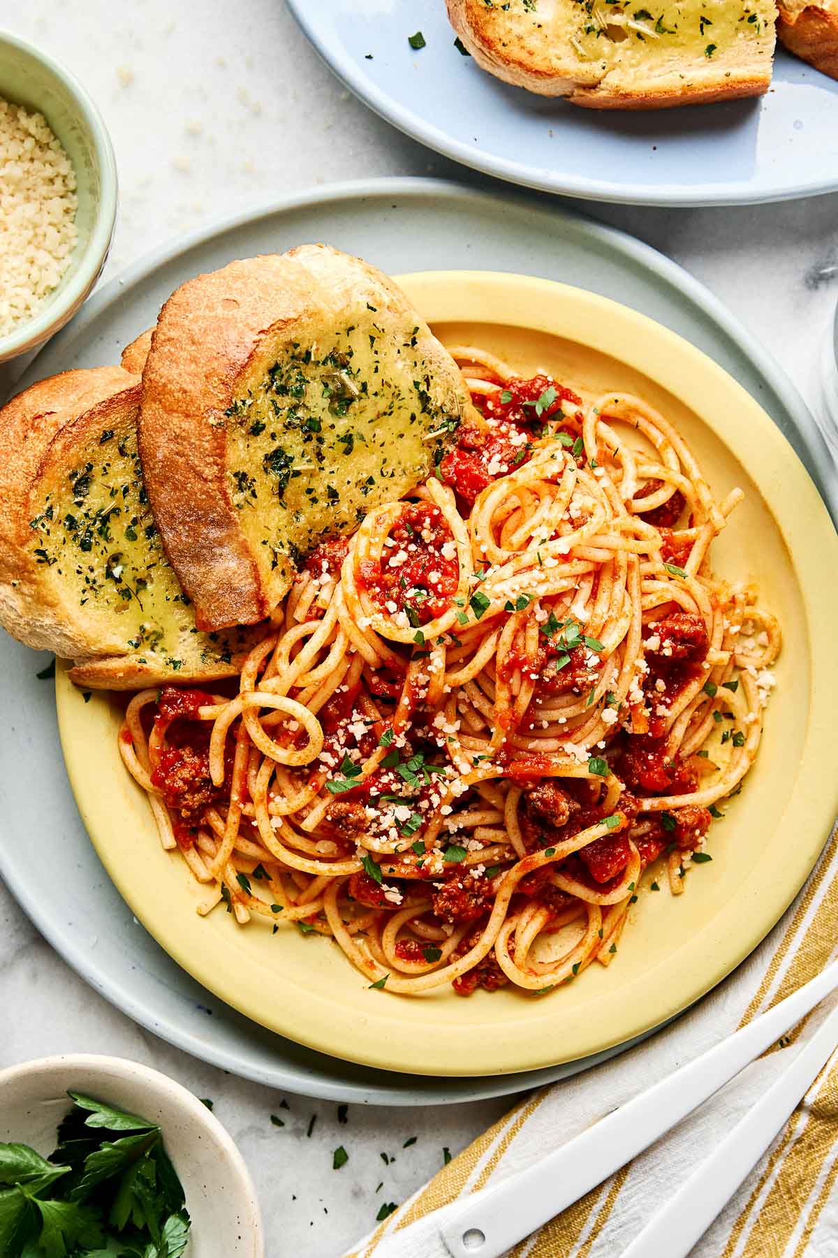 A plate of spaghetti with tomato sauce, topped with grated cheese and chopped herbs, served with two slices of garlic bread on the side. Small bowls of herbs and cheese are nearby.