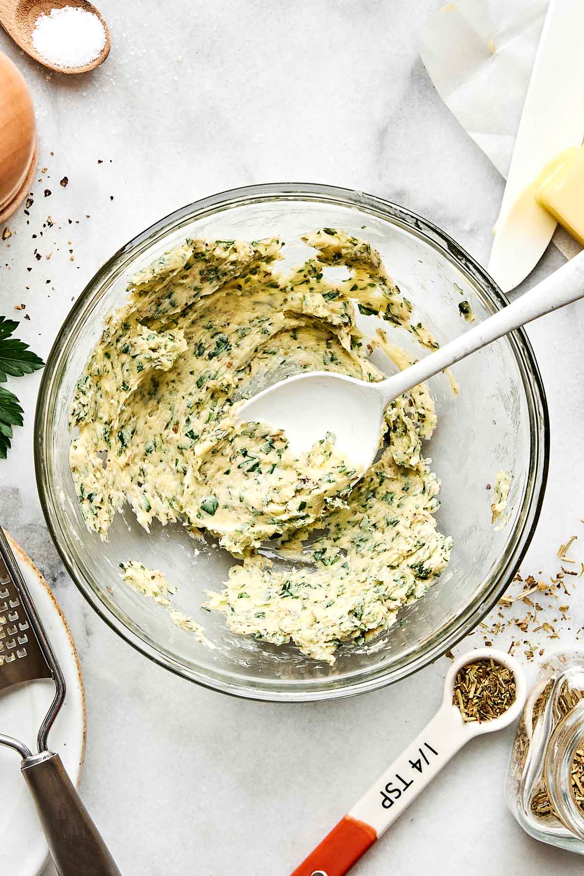A glass bowl with a spoon contains a creamy mixture of butter and chopped herbs. Measuring spoons, a grater, and other cooking utensils are arranged on a white countertop nearby.