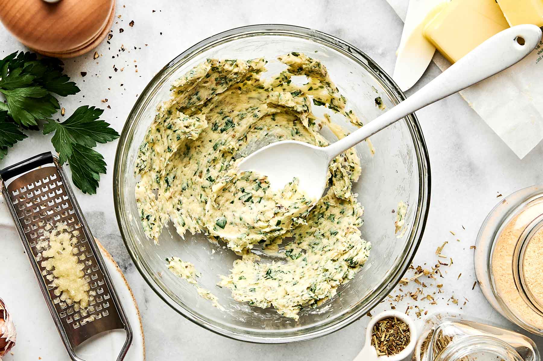 A glass bowl with herb butter and a white spoon, surrounded by a cheese grater with grated garlic, fresh parsley, a butter stick, seasonings, and a jar of breadcrumbs on a white surface.
