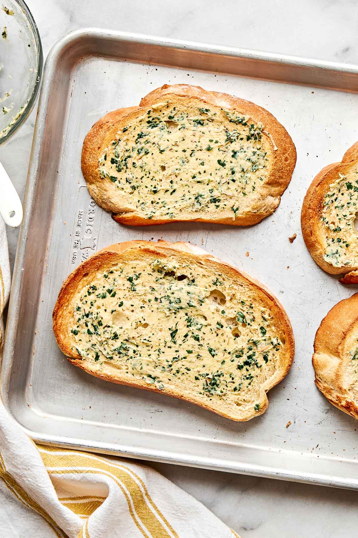 Three slices of toasted bread with a spread of garlic herb butter on a baking sheet, next to a white and yellow striped towel.