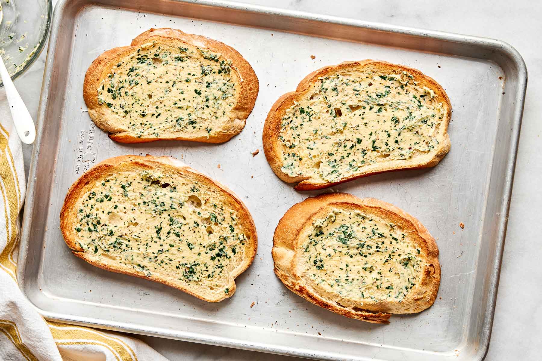 Four slices of toasted bread topped with a garlic and herb spread are arranged on a metal baking sheet. A white towel with yellow stripes is partially visible at the edge of the tray.