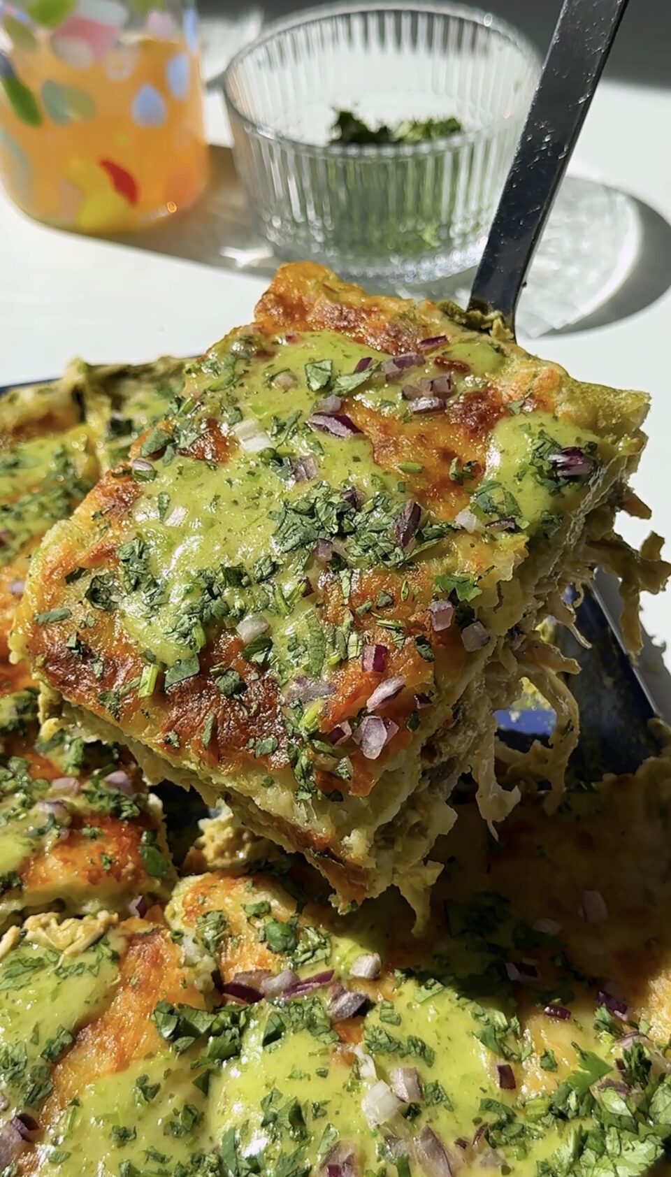 A close-up of a slice of enchiladas being lifted from a pan, topped with melted cheese, chopped herbs, and diced red onions, with a fork visible under the slice in bright natural light.