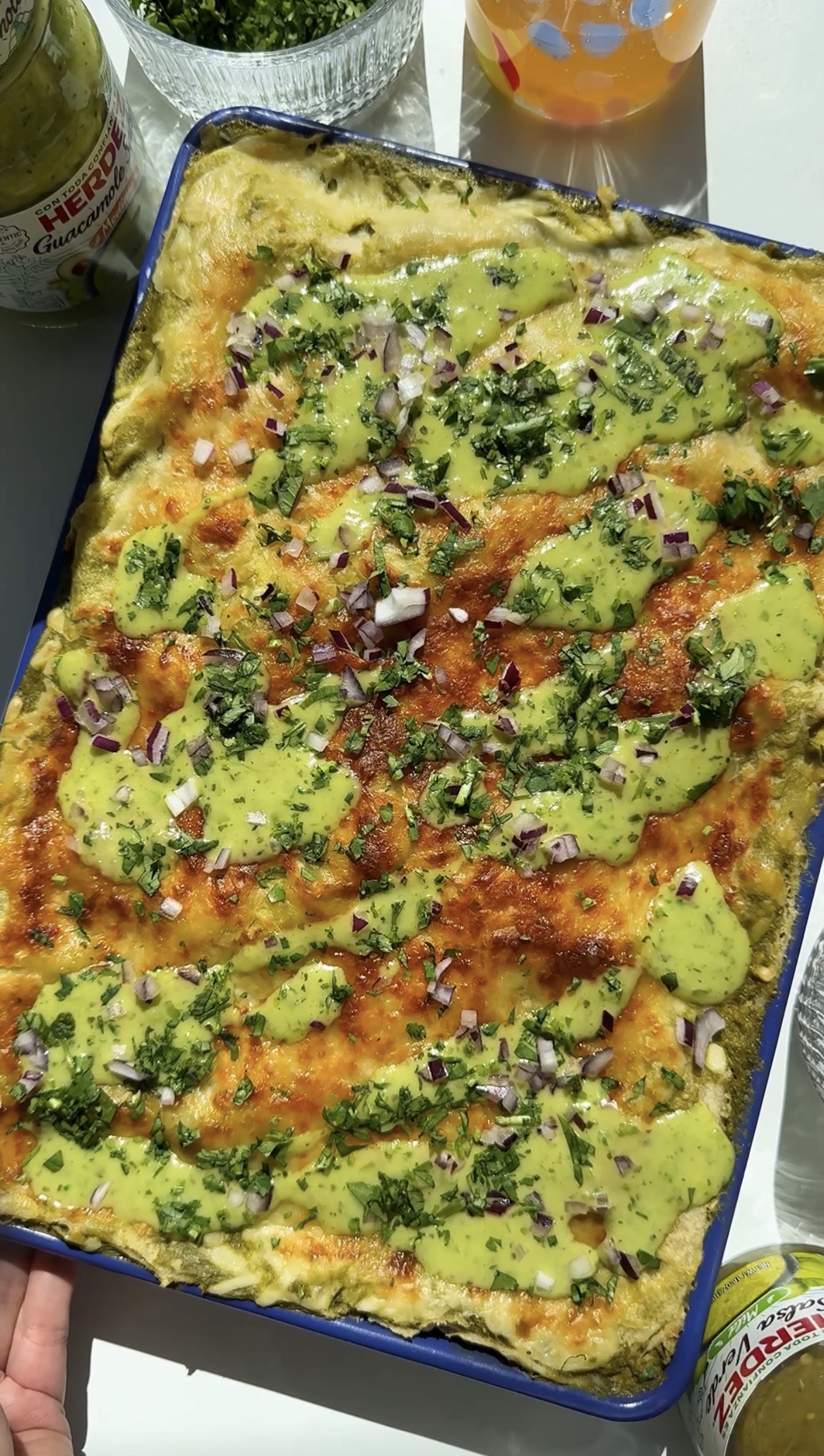 A rectangular pan of enchiladas topped with green sauce, chopped cilantro, diced red onions, and spices, displayed in a pan under bright natural light.