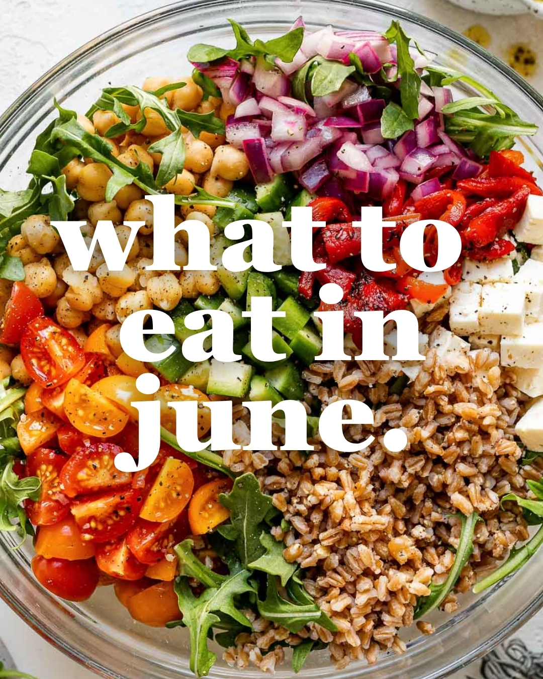 A bowl filled with arugula, chickpeas, chopped red onions, roasted red peppers, feta cheese, cherry tomatoes, farro, and chopped green peppers with the words what to eat in june. overlaid in large white text.