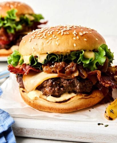 A close-up of a cheeseburger with a sesame seed bun, melted cheese, crispy bacon, lettuce, caramelized onions, and sauce, with another burger blurred in the background.