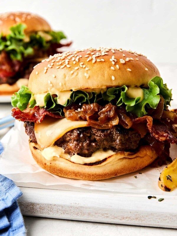 A close-up of a cheeseburger with a sesame seed bun, melted cheese, crispy bacon, lettuce, caramelized onions, and sauce, with another burger blurred in the background.