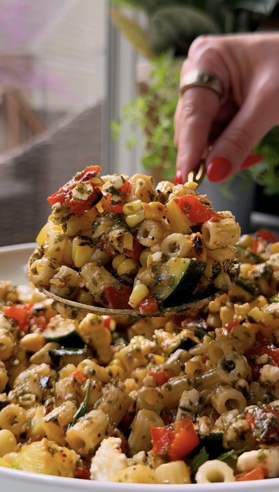 A close-up of a spoon scooping pasta salad with vegetables such as zucchini, corn, and red peppers, mixed with herbs. A hand with red-painted nails holds the spoon over a large serving dish.