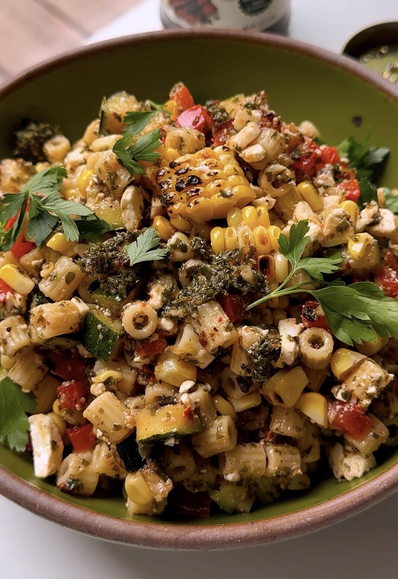A bowl filled with grilled corn salad mixed with chopped vegetables, herbs, and a green dressing, garnished with fresh parsley leaves. The salad appears fresh and colorful.