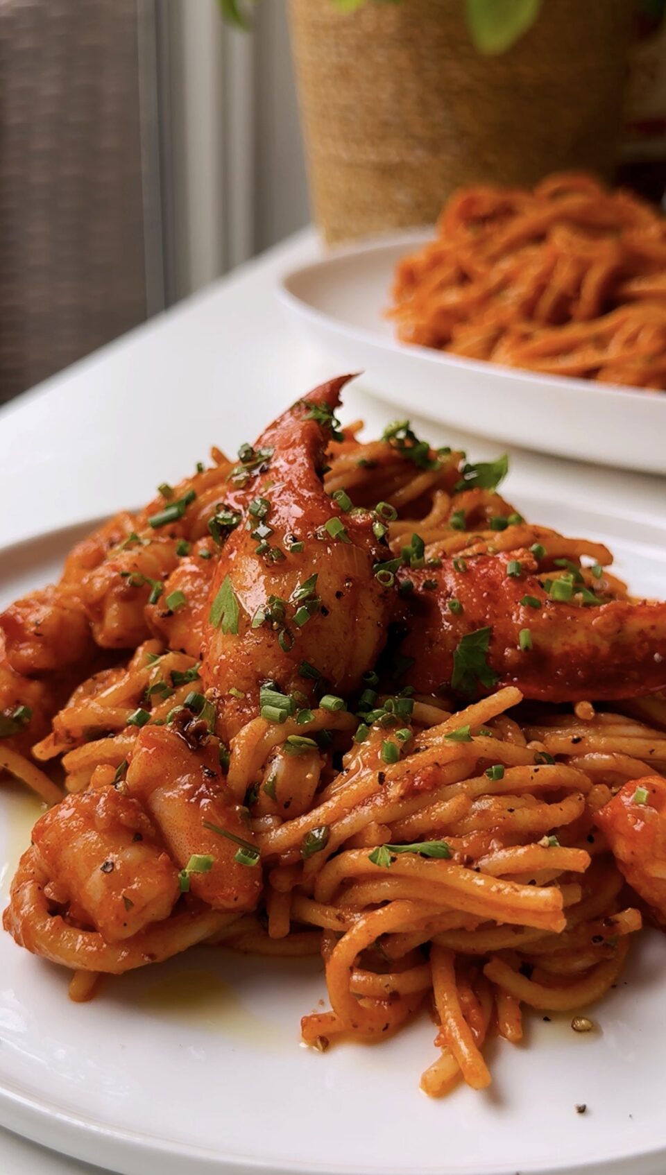 A close-up of spaghetti in a rich red sauce, topped with shrimp, fresh herbs, and black pepper, served on a white plate with another plate of pasta in the background.