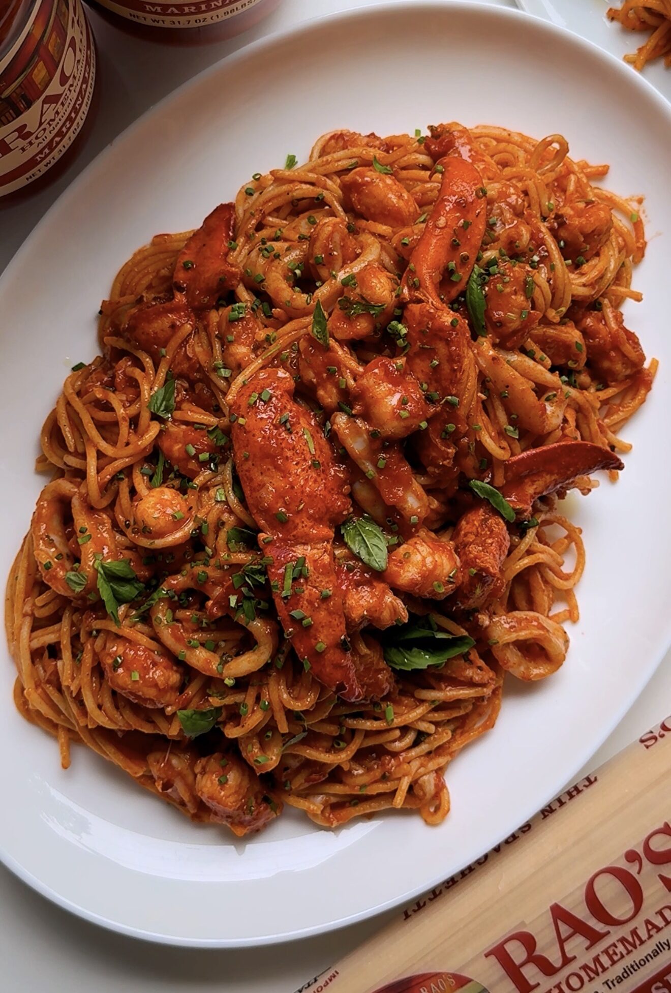 A white oval plate filled with spaghetti and lobster in a rich red tomato sauce, garnished with chopped herbs. Jars of Rao’s Homemade tomato sauce and a wooden utensil are nearby on the white surface.