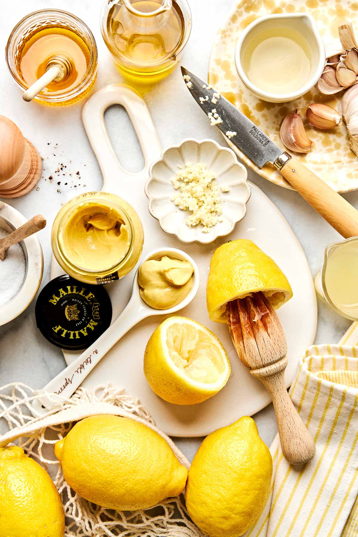 Overhead view of ingredients for a vinaigrette: whole and halved lemons, lemon juice, Dijon mustard, honey, garlic, olive oil, pepper, salt, and kitchen utensils on a white surface with a yellow-striped towel.