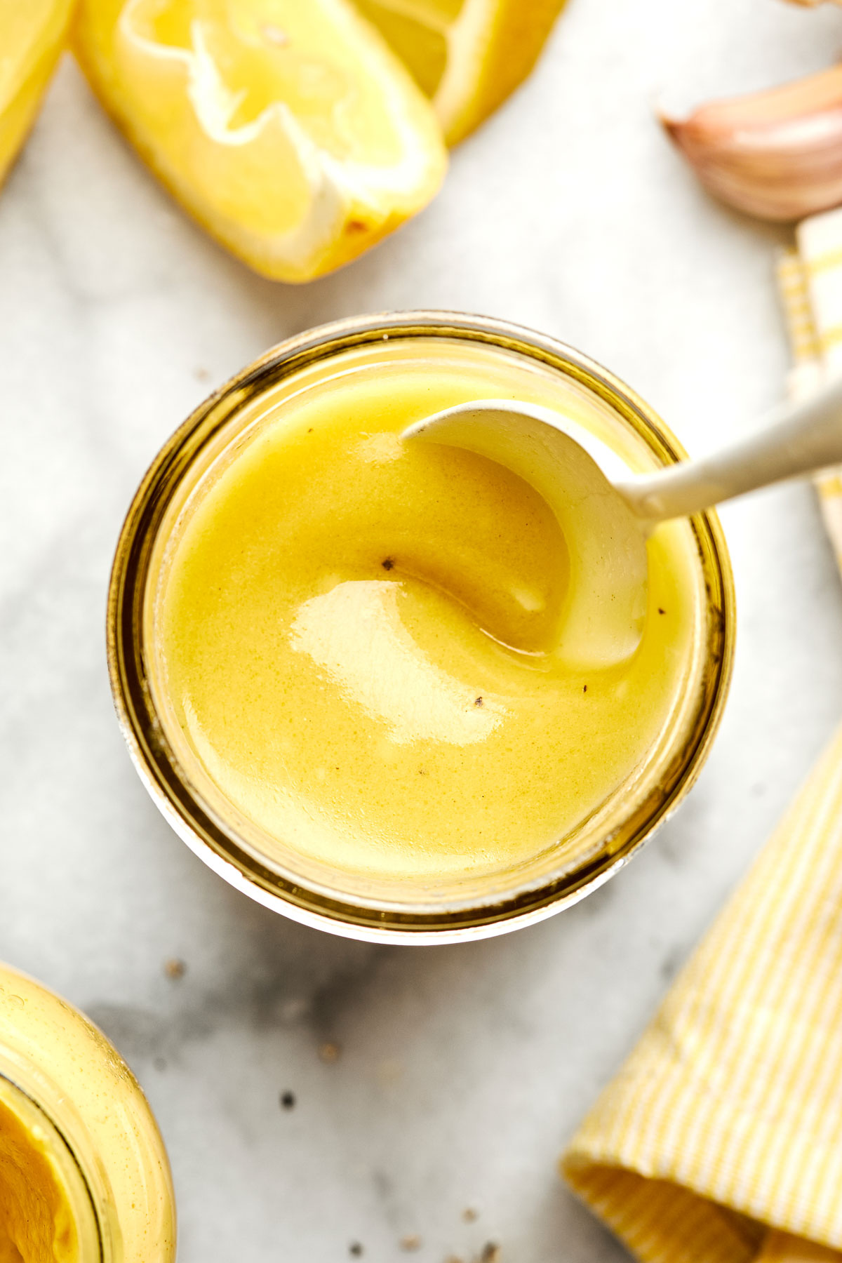 A jar of creamy yellow salad dressing with a spoon, surrounded by lemon wedges, garlic, and a yellow-striped cloth on a marble surface.
