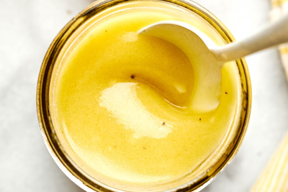 A close-up of a jar filled with creamy yellow honey mustard dressing, with a spoon partially submerged, against a light background.