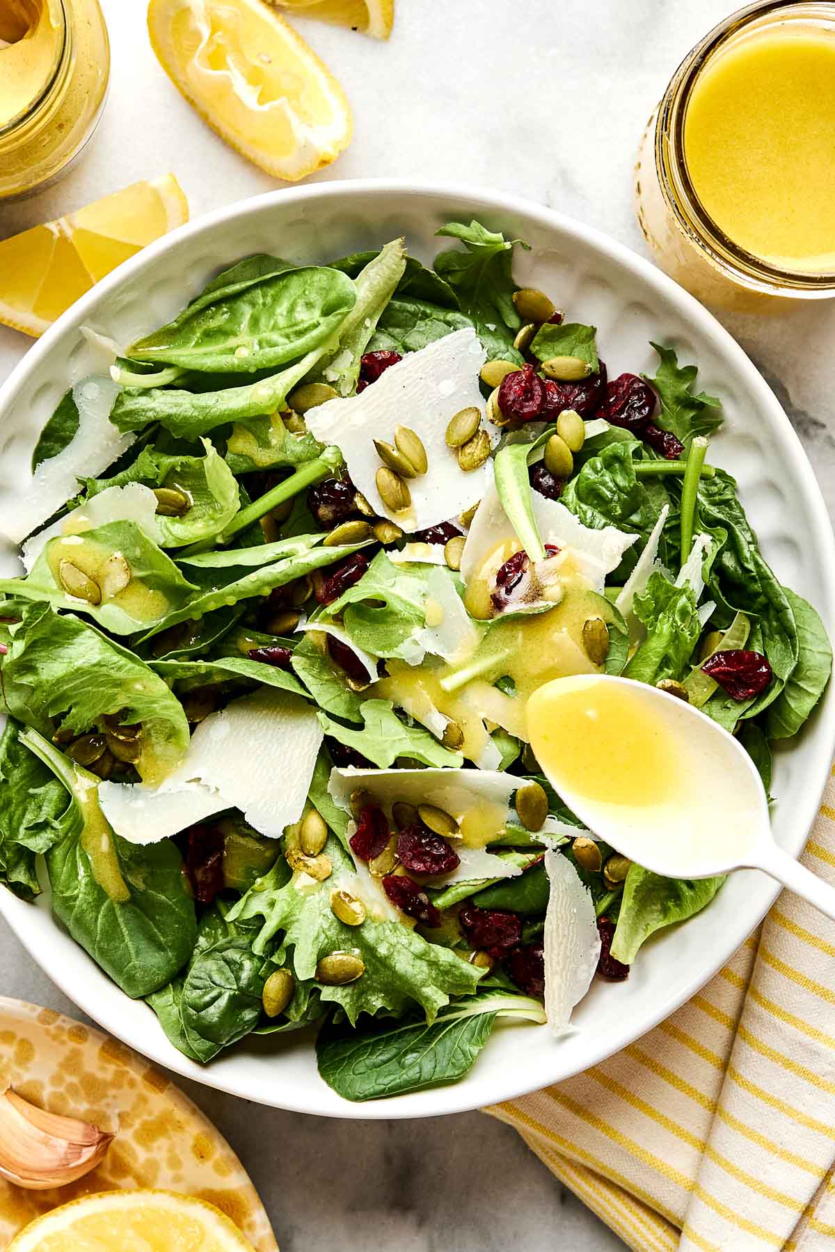 A fresh green salad with spinach, leafy greens, shaved parmesan, pumpkin seeds, dried cranberries, and a lemon vinaigrette being drizzled from a spoon, served in a white bowl. Lemon wedges and dressing are nearby.