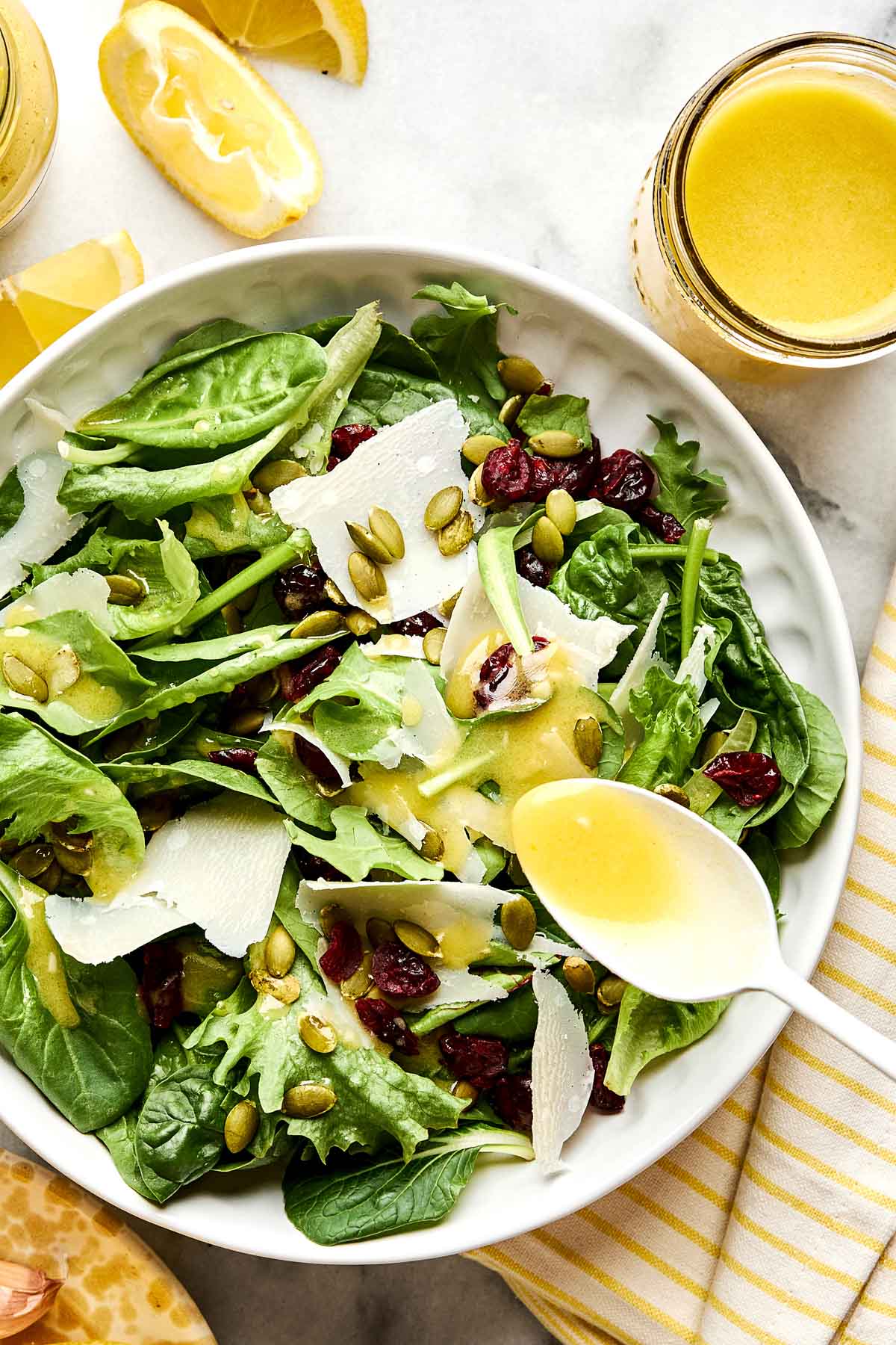 A bowl of mixed green salad with spinach, dried cranberries, pumpkin seeds, and shaved cheese, topped with vinaigrette. A spoon pours more dressing on the salad. Lemon wedges and a jar of dressing are nearby.