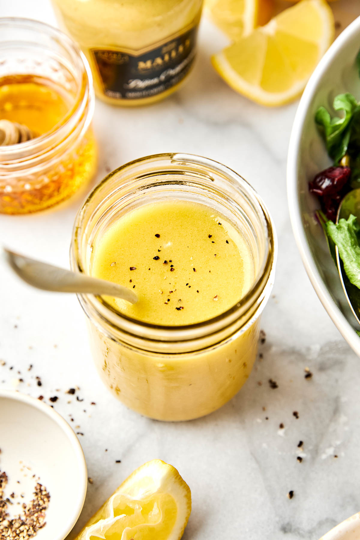 A jar of creamy lemon vinaigrette sits on a marble surface. Nearby are a honey jar, a mustard jar, lemon wedges, black pepper, and a bowl of mixed greens. A spoon is in the jar.