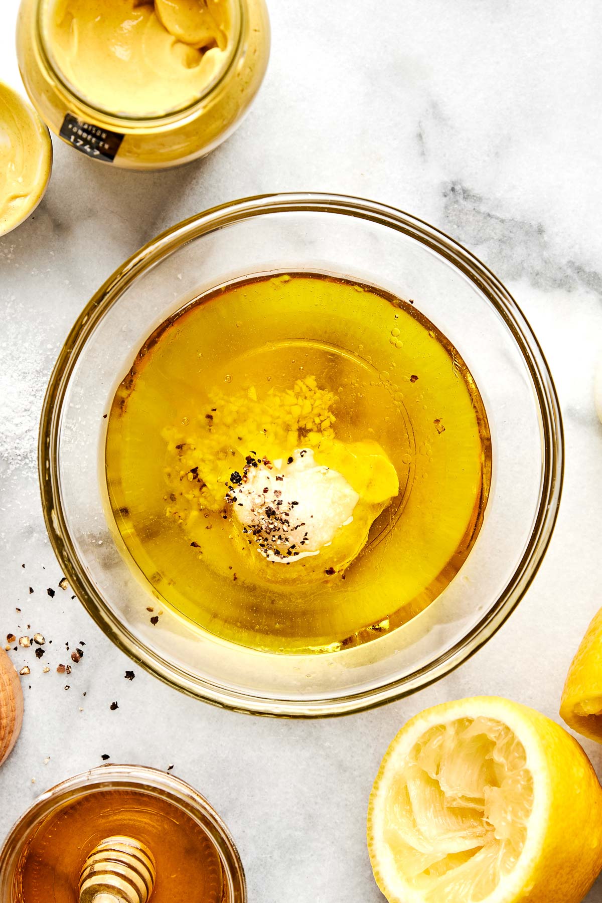 A glass bowl filled with olive oil, minced garlic, mustard, black pepper, and other ingredients sits on a marble surface, surrounded by a halved lemon, a jar of mustard, and a container of honey.