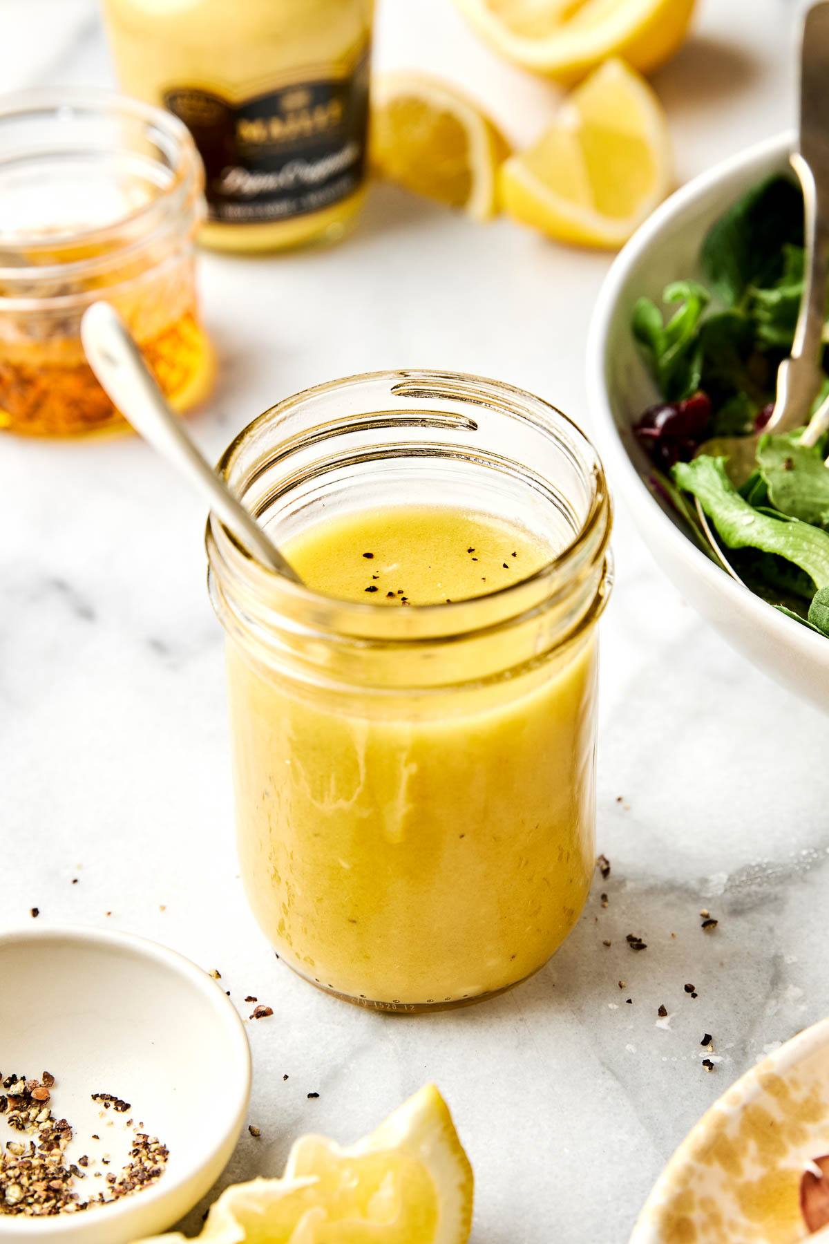 A glass jar filled with yellow salad dressing, topped with cracked black pepper and a spoon inside. Surrounding the jar are lemon wedges, a bowl of greens, and small dishes with pepper and dressing ingredients.