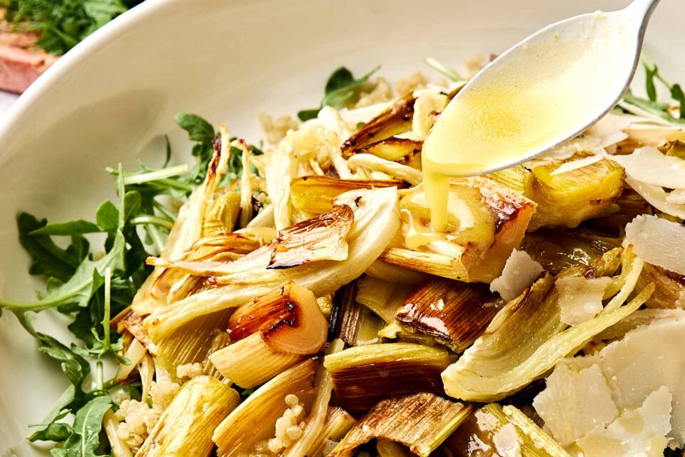 A spoon drizzles dressing over a salad with roasted leeks and fennel, shaved Parmesan cheese, and arugula.
