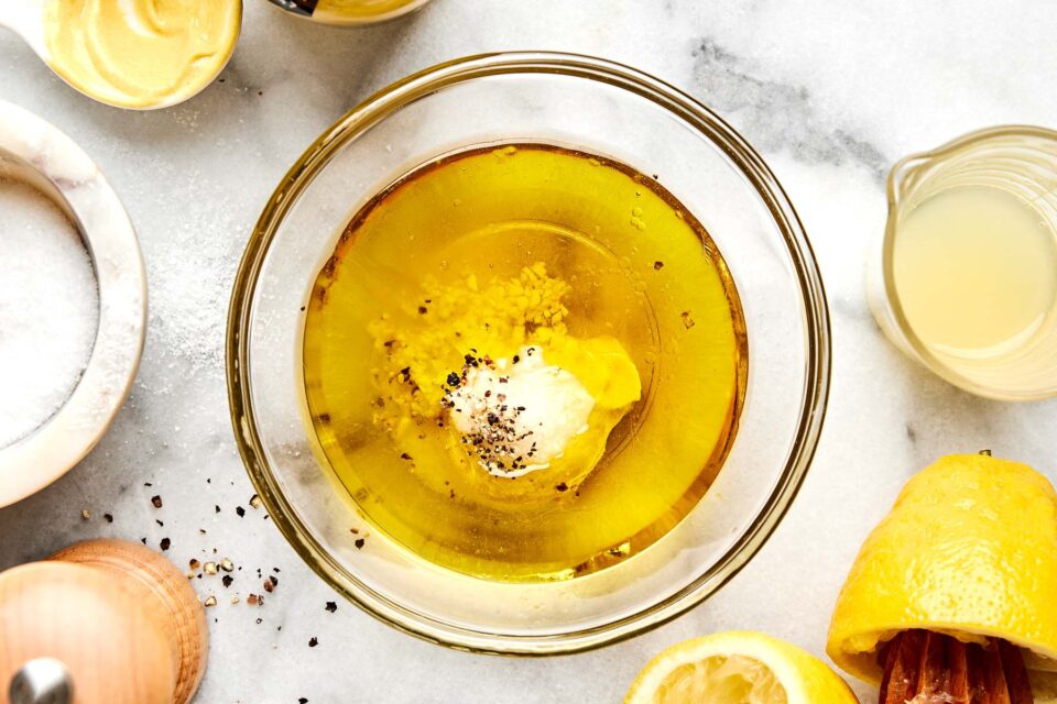 A glass bowl filled with olive oil, minced garlic, mustard, salt, pepper, and lemon juice sits on a marble surface surrounded by a lemon half, a small bowl of mustard, and a cup of lemon juice.