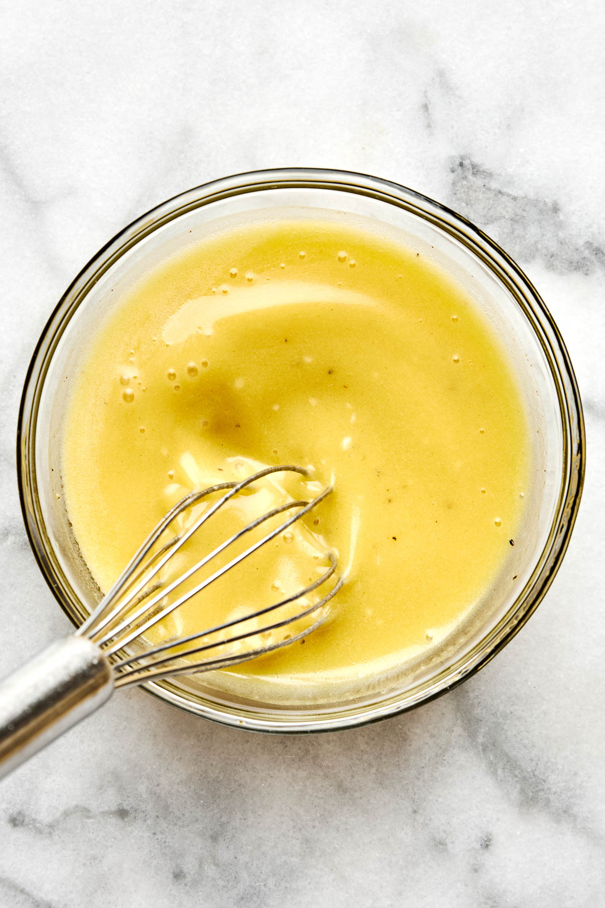 A metal whisk mixing a smooth, pale yellow sauce or batter in a clear glass bowl on a light marble surface.