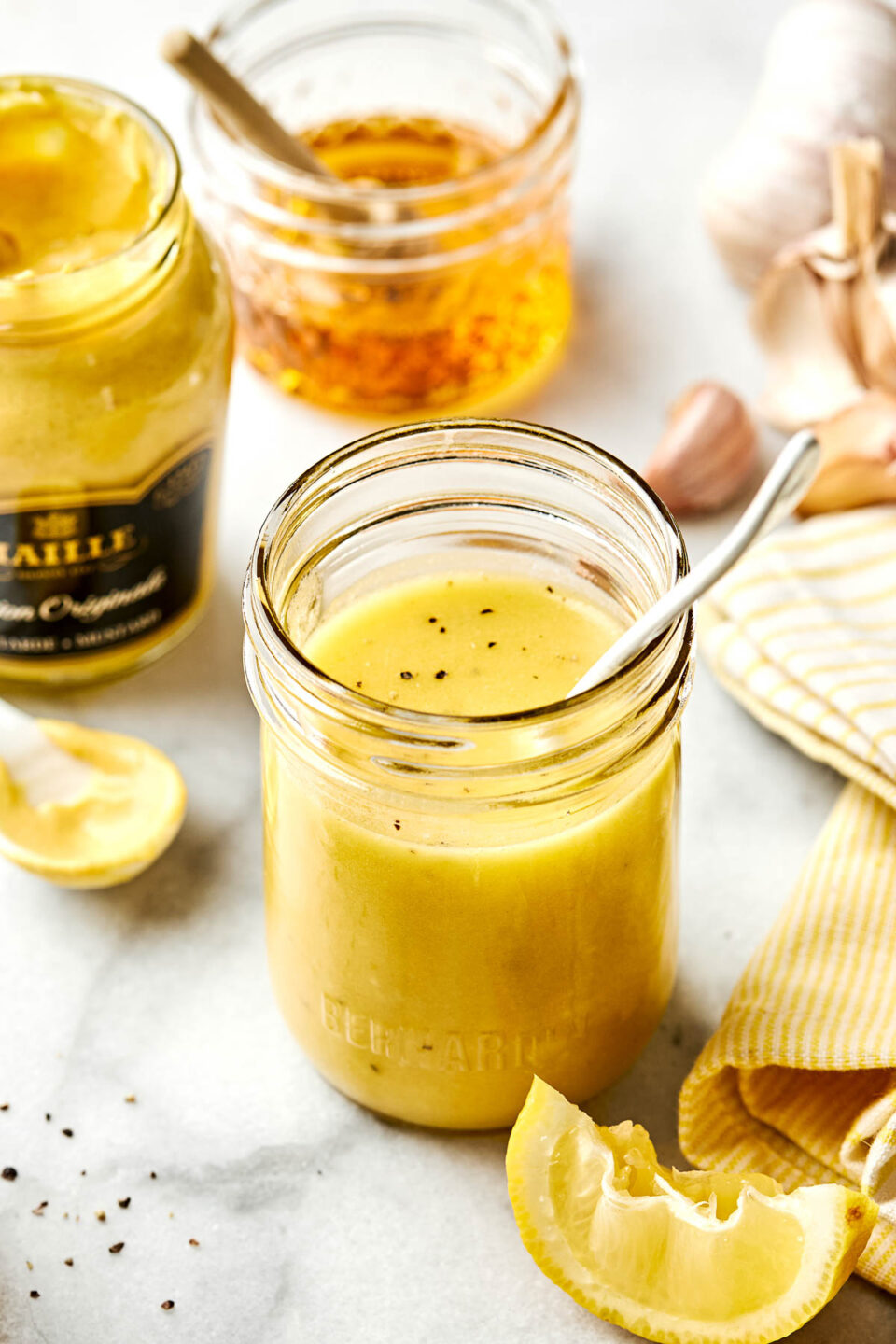 A glass jar filled with creamy yellow dressing and a spoon, surrounded by a mustard jar, honey, garlic cloves, black pepper, a lemon wedge, and a yellow-striped cloth on a white surface.